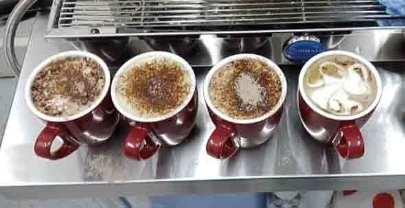 Four mugs of coffees with foam on top on a steel bench. 